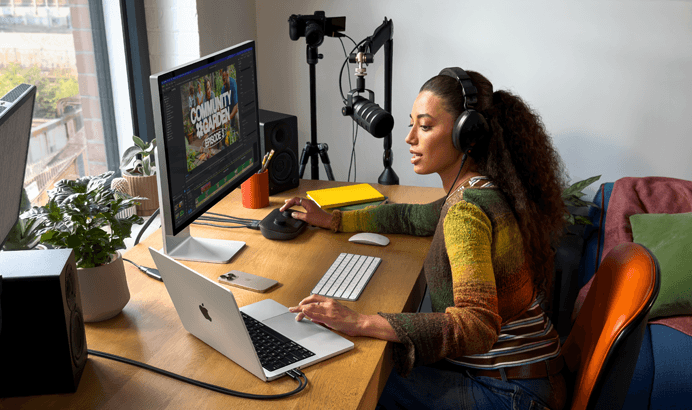 Eine Frau arbeitet bei sich zu Hause an ihrem Schreibtisch mit einem MacBook Pro, das an zwei externe Displays angeschlossen ist. Sie trägt Kopfhörer und spricht in ein Mikrofon